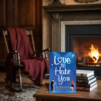 A book titled "Love to Hate You" by Jo Watson stands upright on a wooden table in a cozy room with a burning fireplace, a rocking chair draped with a red blanket, and a stack of books nearby.