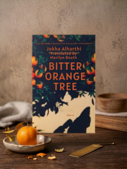 Book titled 'Bitter Orange Tree' by Jokha Alharthi on a wooden surface with an orange and a bowl.